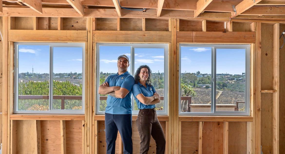 Two people standing in a room with exposed wooden studs and new windows.